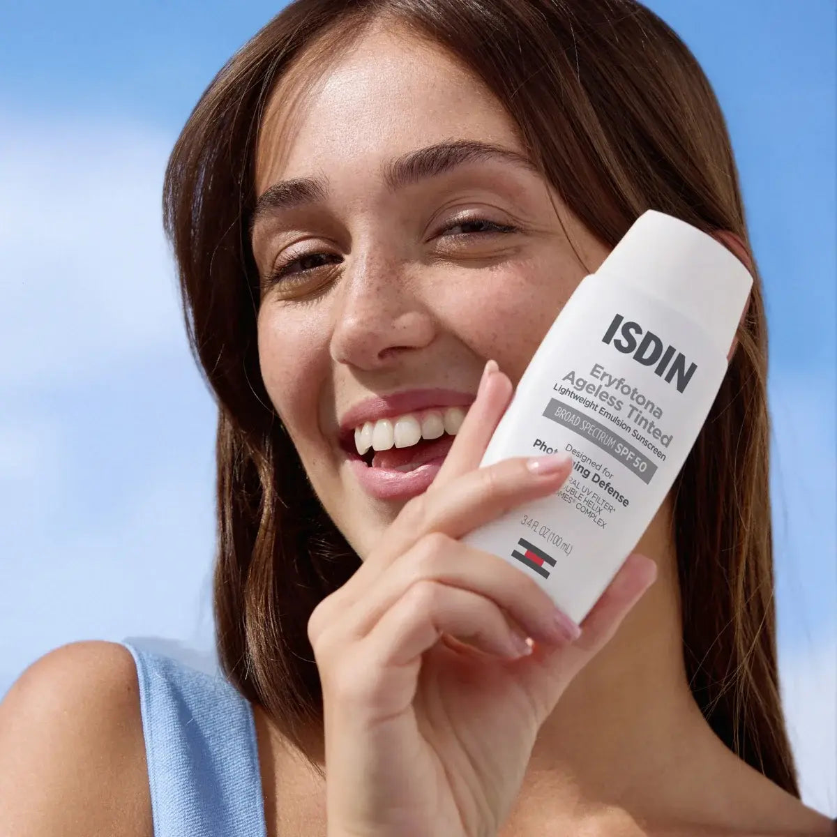 Smiling brunette woman with freckles holding a bottle of ISDIN Eryfotona Ageless Tinted SPF 50 against a bright blue sky. 