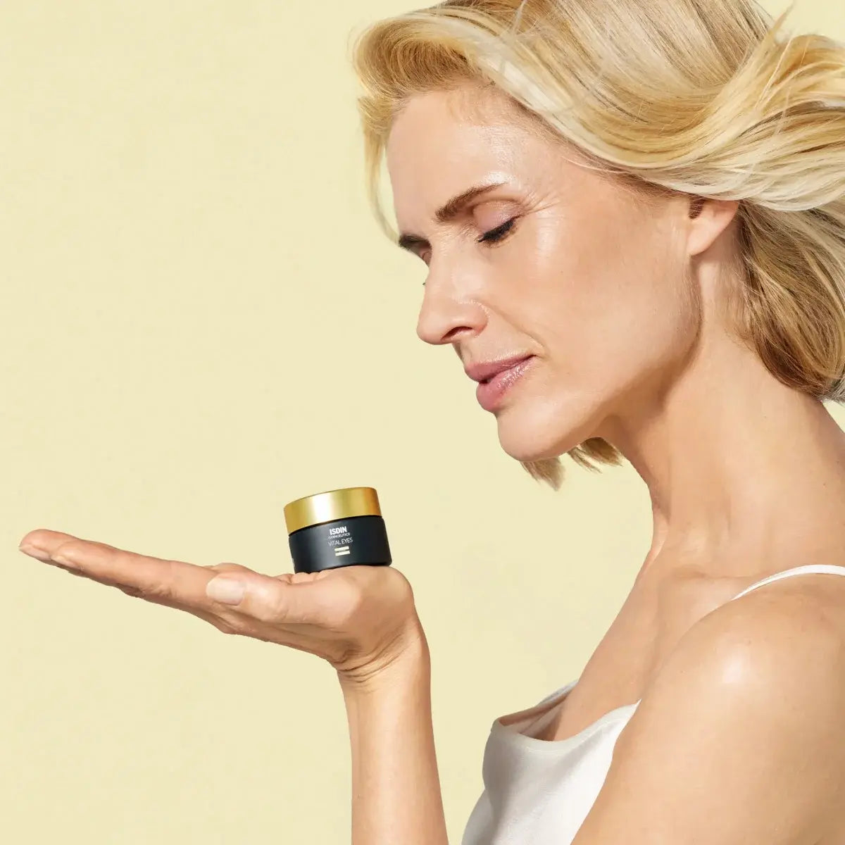 Middle aged woman with healthy-looking skin holding a canister of ISDINCEUTICS Vital Eyes in her palm against a pale yellow background.