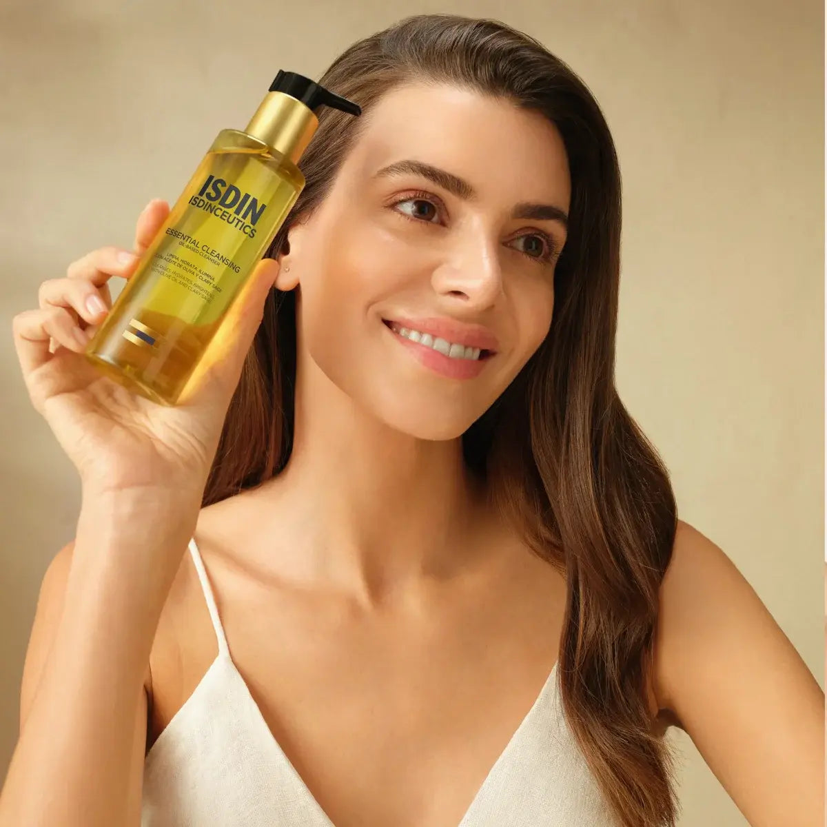 Woman with healthy-looking skin holding a golden pump bottle of ISDINCEUTICS Essential Cleansing Oil near her head against a beige background. 