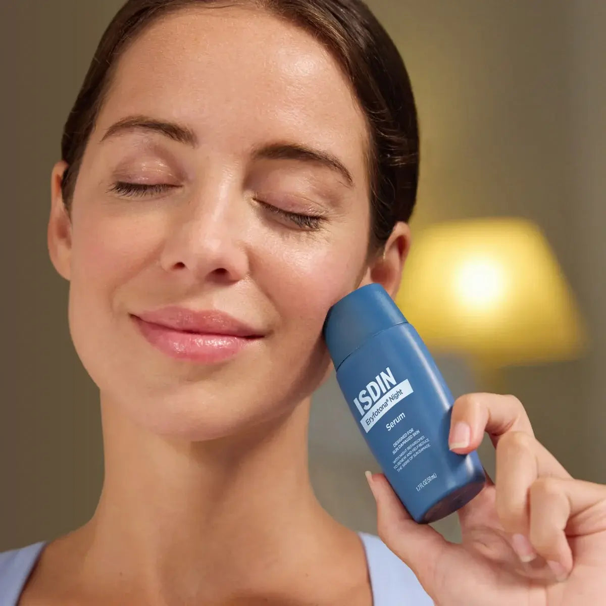 Close up of woman with healthy-looking skin holding a blue bottle of ISDIN Eryfotona Night Serum near her face. 